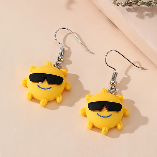 A pair of yellow sun-shaped dangle earrings with black sunglasses and blue smiling mouths, hanging from silver fishhook wires on a light peach background.